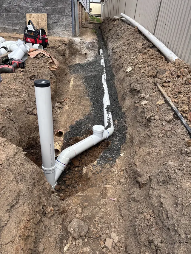 New PVC drainage pipe laid on gravel in an open trench beside a house