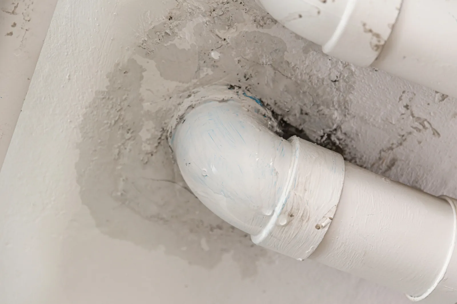 Water droplets on a leaking white pipe elbow with a spreading damp stain on the plaster around it